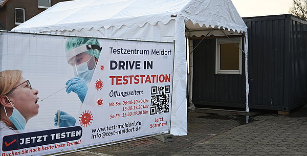 Ein Banner der Drive-In Teststation, das vor einem weißen Zelt steht. Dort fahren die Autos durch